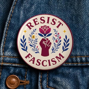 Könnte beinhalten: Ein runder Button mit dem Schriftzug "RESIST FASCISM" in Burgunderrot. Der Button zeigt eine burgunderrote Faust, die eine Blume hält, umgeben von floralen und Blattdesigns in Blau, Gelb und Rosa, vor einem cremefarbenen Hintergrund.