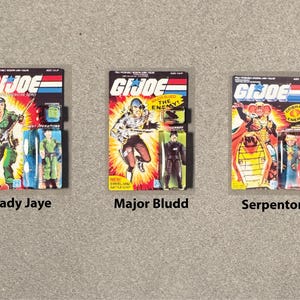 May include: Three G.I. Joe action figure packages. Lady Jaye, in green, holds a rifle. Major Bludd, in brown, is mid-action. Serpentor, in yellow and red, is on a throne. Each package has the G.I. Joe logo.