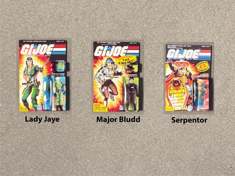 May include: Three G.I. Joe action figure packages. Lady Jaye, in green, holds a rifle. Major Bludd, in brown, is mid-action. Serpentor, in yellow and red, is on a throne. Each package has the G.I. Joe logo.