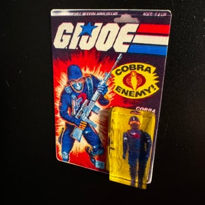 May include: A vintage G.I. Joe action figure card and a small, sealed figure. The card features a blue-clad soldier with a weapon, the G.I. Joe logo, and the text "COBRA ENEMY!". The small figure is in a yellow plastic case.