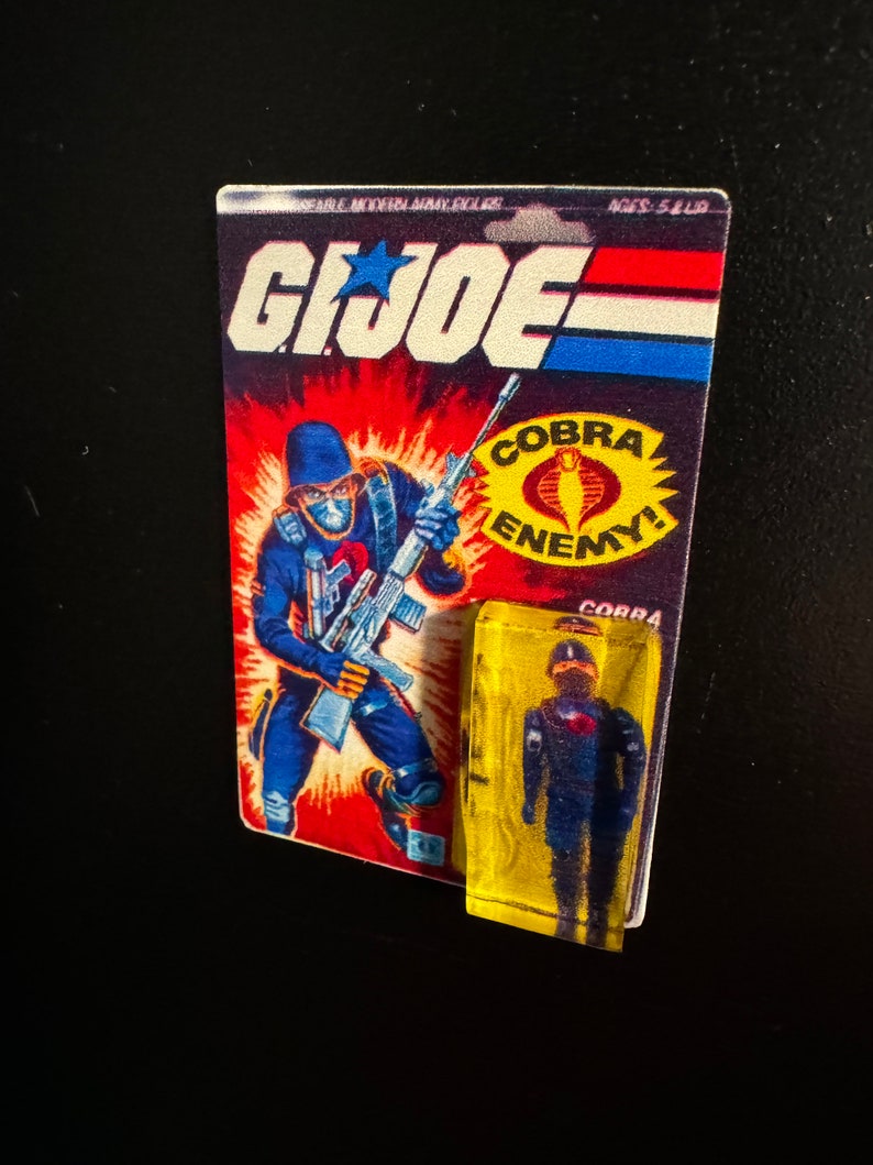 May include: A vintage G.I. Joe action figure card and a small, sealed figure. The card features a blue-clad soldier with a weapon, the G.I. Joe logo, and the text "COBRA ENEMY!". The small figure is in a yellow plastic case.