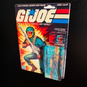 May include: A vintage G.I. Joe action figure on its original card. The card features a soldier illustration with a machine gun against a fiery background. The packaging includes the text "G.I. Joe A Real American Hero" and "Gunner".