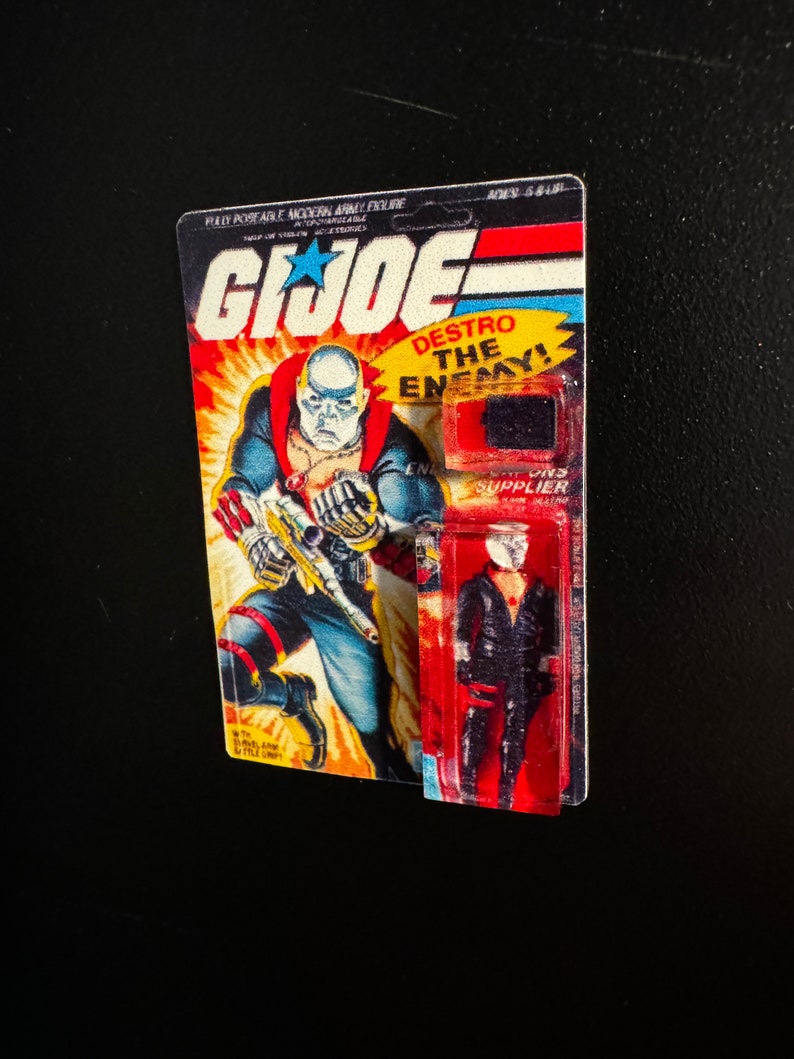 May include: Vintage G.I. Joe action figure on its original card. The card features a comic book style illustration of a bald character in a blue and red uniform. The text "DESTRO THE ENEMY!" is visible. The figure is in a clear plastic bubble.