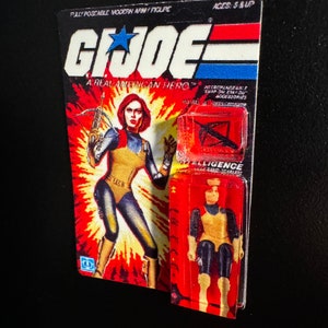 May include: A vintage G.I. Joe action figure on its original card. The card features a female character with red hair, wearing a yellow and blue outfit, and holding a crossbow. The packaging reads "G.I. Joe: A Real American Hero."