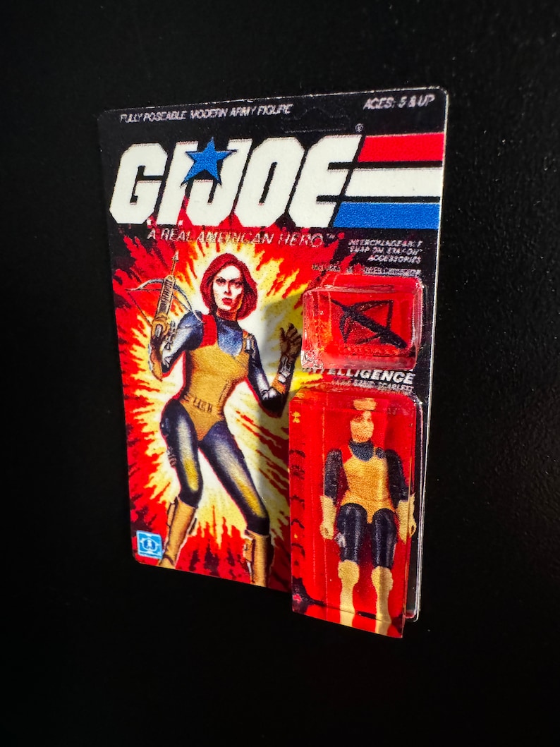 May include: A vintage G.I. Joe action figure on its original card. The card features a female character with red hair, wearing a yellow and blue outfit, and holding a crossbow. The packaging reads "G.I. Joe: A Real American Hero."
