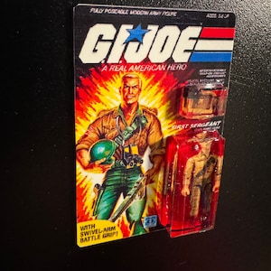 May include: A vintage G.I. Joe action figure, still in its original packaging. The card features a muscular soldier illustration against a fiery background. The packaging includes the text "A Real American Hero" and "First Sergeant".