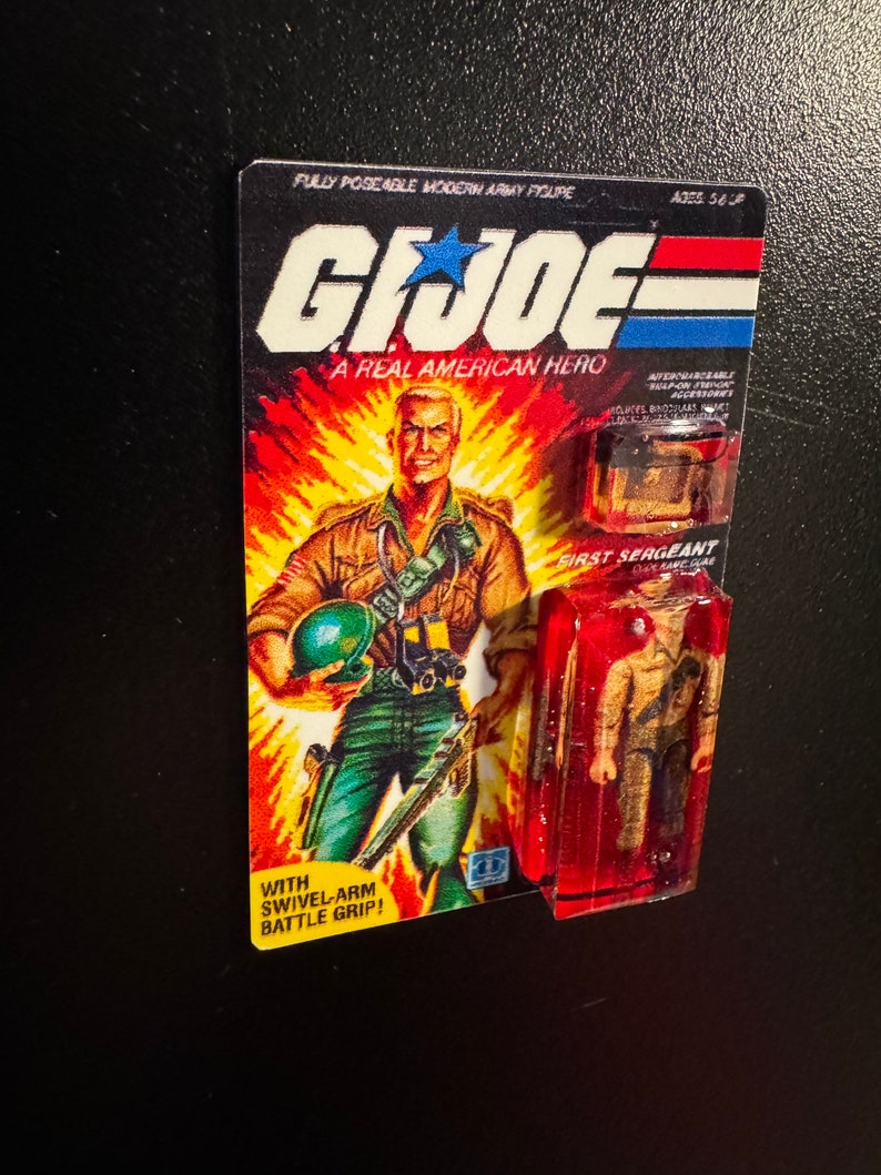 May include: A vintage G.I. Joe action figure, still in its original packaging. The card features a muscular soldier illustration against a fiery background. The packaging includes the text "A Real American Hero" and "First Sergeant".