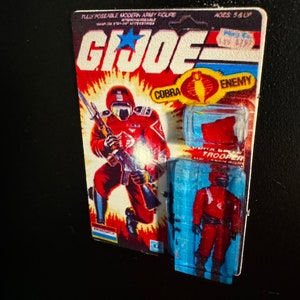 May include: A vintage G.I. Joe Cobra Trooper action figure on its original card. The card features a red and orange background with the G.I. Joe logo and a Cobra symbol. The figure is red and black.
