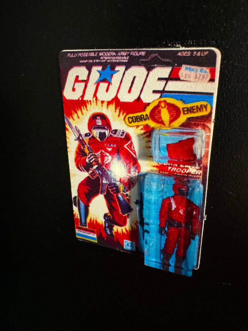 May include: A vintage G.I. Joe Cobra Trooper action figure on its original card. The card features a red and orange background with the G.I. Joe logo and a Cobra symbol. The figure is red and black.