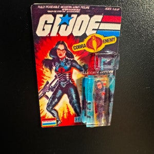 May include: A vintage G.I. Joe action figure card featuring a female character in a black and blue outfit, holding a rifle. The card reads "G.I. Joe" with "COBRA ENEMY" and "INTELLIGENCE OFFICER". The figure is in a clear plastic case.