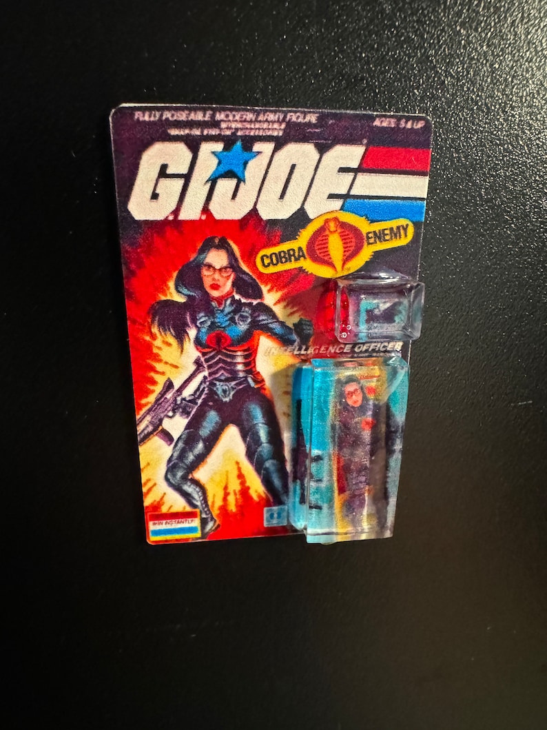 May include: A vintage G.I. Joe action figure card featuring a female character in a black and blue outfit, holding a rifle. The card reads "G.I. Joe" with "COBRA ENEMY" and "INTELLIGENCE OFFICER". The figure is in a clear plastic case.