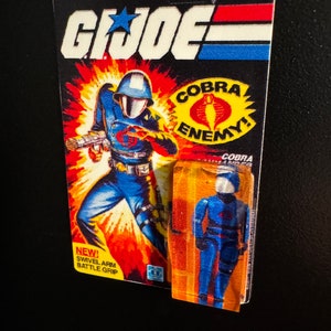 May include: A vintage G.I. Joe action figure on its original packaging. The packaging features a dynamic illustration of a soldier in blue, with the words "COBRA ENEMY!" and "SWIVEL ARM BATTLE GRIP". The figure is fully poseable.