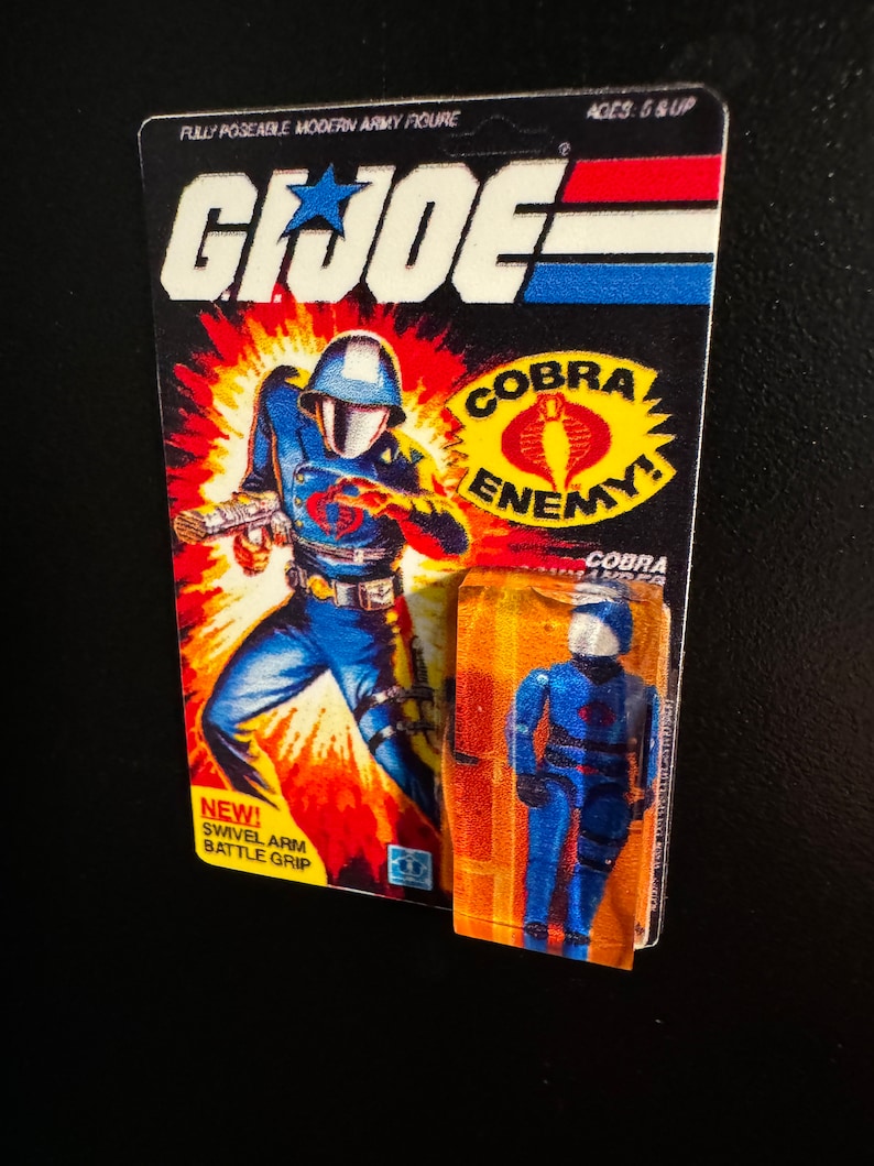 May include: A vintage G.I. Joe action figure on its original packaging. The packaging features a dynamic illustration of a soldier in blue, with the words "COBRA ENEMY!" and "SWIVEL ARM BATTLE GRIP". The figure is fully poseable.