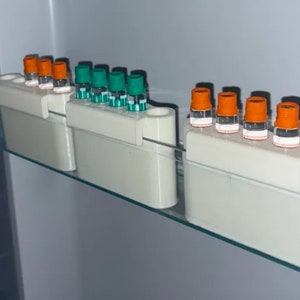 Op de afbeelding: Verschillende kleine glazen flesjes met oranje en groene doppen zijn gerangschikt in witte houders in een koelkast. De flesjes zijn gevuld met een heldere vloeistof. De houders zijn rechthoekig.