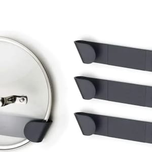 Puede incluir: Tres soportes de metal negro para tapas de ollas con una tapa de olla plateada colgada en uno de ellos. Los soportes están diseñados para montarse en la pared.