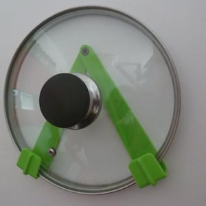 May include: A clear glass pot lid with a stainless steel rim and a black handle. The lid is held in place by a bright green plastic holder. The holder is attached to the wall.
