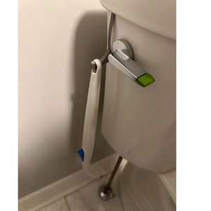 Può includere: Uno spazzolino per WC bianco con un manico blu e verde è appeso a un serbatoio del WC bianco. Lo spazzolino è attaccato a un gancio argentato con un pulsante verde.