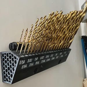 May include: A black drill bit holder with a honeycomb design on the side, mounted on a wall. The holder is filled with numerous gold-colored drill bits of varying sizes, labeled with fractional inch measurements.