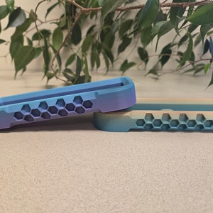 Puede incluir: Dos fundas para teléfono impresas en 3D con un diseño de panal. La funda superior es azul y morada, mientras que la funda inferior es beige y azul.