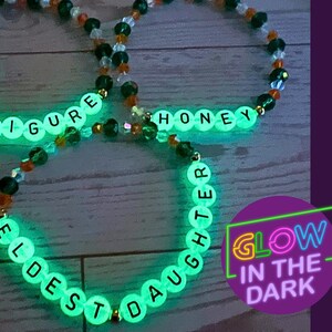 Puede incluir: Tres pulseras de cuentas que brillan en la oscuridad. Una pulsera deletrea "FATHER FIGURE", otra "HONEY" y la tercera "ELDEST DAUGHTER". Las pulseras tienen cuentas transparentes con cuentas de letras blancas y detalles de cuentas naranjas y negras. Una pegatina morada dice "GLOW IN THE DARK".