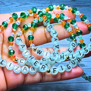 Puede incluir: Varios brazaletes de cuentas con cuentas verdes, naranjas y transparentes. Algunos brazaletes tienen cuentas de letras blancas que deletrean palabras como "ELIZABETH", "FATHER", "WISHED" y "FIGURE". Los brazaletes se sostienen en una mano.