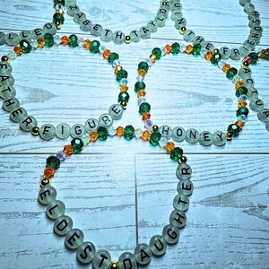Puede incluir: Varios collares de cuentas con palabras como "FATHER" y "HONEY". Las cuentas verde claro tienen letras negras, intercaladas con cuentas de cristal verde, naranja y transparente. Tambi&eacute;n hay cuentas doradas.