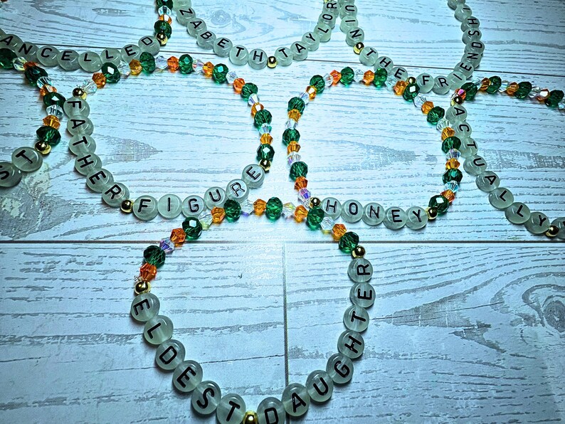 Puede incluir: Varios collares de cuentas con palabras como "FATHER" y "HONEY". Las cuentas verde claro tienen letras negras, intercaladas con cuentas de cristal verde, naranja y transparente. Tambi&eacute;n hay cuentas doradas.