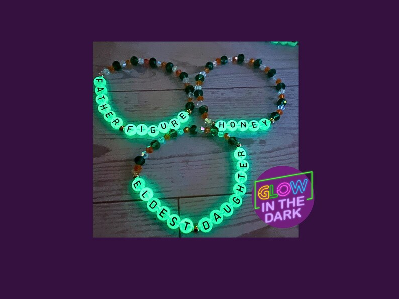 Puede incluir: Tres pulseras de cuentas que brillan en la oscuridad. Una pulsera deletrea "FATHER FIGURE", otra "HONEY" y la tercera "ELDEST DAUGHTER". Las pulseras tienen cuentas transparentes con cuentas de letras blancas y detalles de cuentas naranjas y negras. Una pegatina morada dice "GLOW IN THE DARK".
