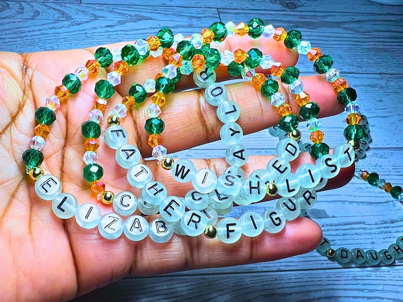 Puede incluir: Varios brazaletes de cuentas con cuentas verdes, naranjas y transparentes. Algunos brazaletes tienen cuentas de letras blancas que deletrean palabras como "ELIZABETH", "FATHER", "WISHED" y "FIGURE". Los brazaletes se sostienen en una mano.
