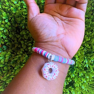 May include: A colorful bracelet featuring a white donut charm with pink, green, and blue sprinkles. The bracelet is made of small, flat, round beads in shades of pink, white, blue, and gray. The charm hangs from a silver-colored chain.