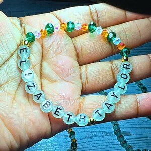Puede incluir: Una pulsera de cuentas con los nombres "ELIZABETH" y "TAYLOR" escritos en letras negras sobre cuentas transl&uacute;cidas. La pulsera presenta cuentas de acento verdes, naranjas y transparentes, y espaciadores dorados.