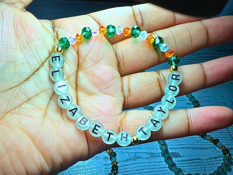 Puede incluir: Una pulsera de cuentas con los nombres "ELIZABETH" y "TAYLOR" escritos en letras negras sobre cuentas transl&uacute;cidas. La pulsera presenta cuentas de acento verdes, naranjas y transparentes, y espaciadores dorados.