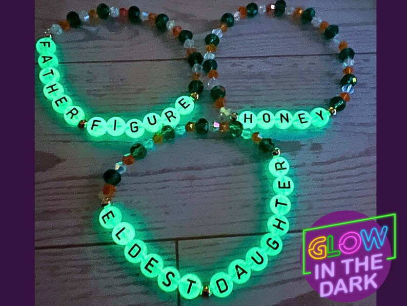Puede incluir: Tres pulseras de cuentas que brillan en la oscuridad. Cada pulsera tiene cuentas de letras blancas que deletrean palabras como "FATHER FIGURE" y "HONEY". Las pulseras est&aacute;n ensartadas con cuentas transparentes, verdes y naranjas. La imagen incluye un letrero de ne&oacute;n que dice "GLOW IN THE DARK".
