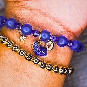 Peut inclure: Deux bracelets sont présentés. Le bracelet supérieur est composé de perles bleues avec des breloques dorées, dont une étoile, un cœur et un croissant de lune. Le bracelet inférieur est composé de perles dorées. Les bracelets sont portés sur un poignet.