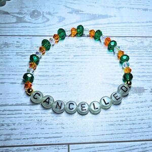 Puede incluir: Una pulsera de cuentas con la palabra "CANCELLED" escrita con cuentas de letras blancas. La pulsera presenta cuentas verdes, naranjas y transparentes, con detalles dorados. La pulsera est&aacute; sobre una superficie de madera blanca y azul claro.