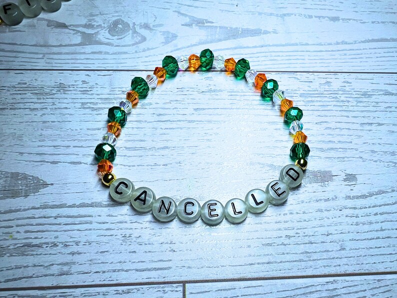 Puede incluir: Una pulsera de cuentas con la palabra "CANCELLED" escrita con cuentas de letras blancas. La pulsera presenta cuentas verdes, naranjas y transparentes, con detalles dorados. La pulsera est&aacute; sobre una superficie de madera blanca y azul claro.