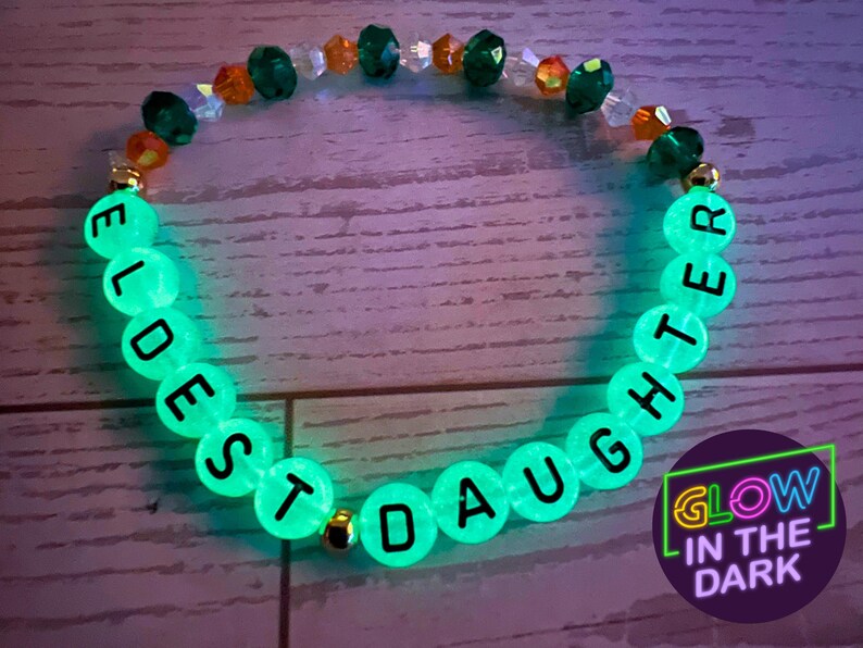 Puede incluir: Pulsera que brilla en la oscuridad con las palabras "ELDEST DAUGHTER" en letras negras sobre cuentas verdes brillantes. La pulsera tambi&eacute;n presenta cuentas doradas y detalles de cuentas naranjas y transparentes. Un c&iacute;rculo p&uacute;rpura con "GLOW IN THE DARK" est&aacute; en la esquina inferior derecha.