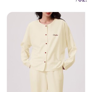 May include: A cream-colored pajama set with a round neckline and batwing sleeves. The top has a button closure and the text "Camellia" embroidered on the right side.