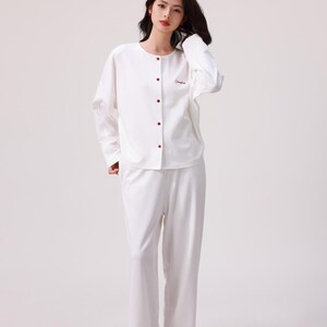 May include: A white long-sleeved pajama top with a button-up front and a small embroidered logo. The matching white pajama pants have a relaxed fit.
