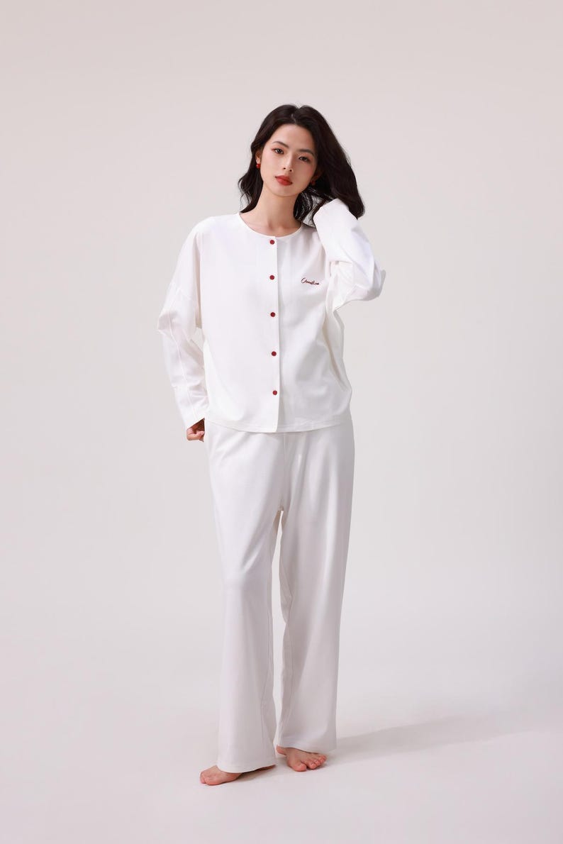 May include: A white long-sleeved pajama top with a button-up front and a small embroidered logo. The matching white pajama pants have a relaxed fit.