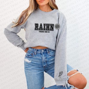 May include: Gray sweatshirt with black text that reads "RAIHN THERE SHE IS". The sweatshirt has a small gray wing design on the left sleeve.