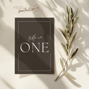 May include: A dark gray table number card with the text "table no. ONE" in white script and a white border. The card is next to a sprig of olive branches.