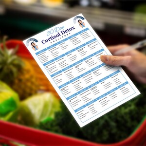 Puede incluir: Una hoja impresa en blanco titulada "30 Day Cortisol Detox Challenge" se sostiene en una mano. La hoja tiene un borde azul y enumera planes de comidas diarios. El fondo muestra productos frescos en un carrito de compras.