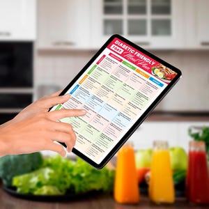 May include: A tablet displaying a 7-day diabetic-friendly meal plan. The screen shows a colorful grid with meal suggestions for breakfast, lunch, snack, and dinner. The tablet is held in front of a blurred background with fresh produce and juice bottles.