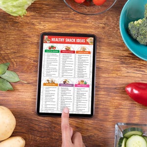 May include: A tablet screen displaying a list of healthy snack ideas, categorized by food type. The categories include fruits & veggies, protein packs, whole grains, healthy fats, nuts & spreads, and sweet treats. The tablet is on a wooden table with various fruits and vegetables around it.