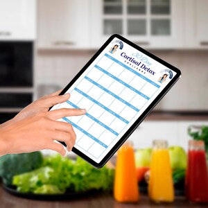 Puede incluir: Una tableta que muestra un calendario del Desaf&iacute;o de Desintoxicaci&oacute;n de Cortisol de 30 d&iacute;as. La cuadr&iacute;cula del calendario es azul claro con texto blanco. La tableta se sostiene en una mano, con un dedo apuntando a la pantalla. Botellas de jugo coloridas y productos frescos en el fondo.