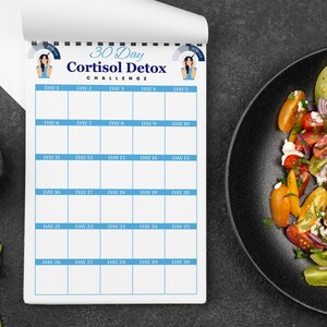 Puede incluir: Un bloc de notas blanco con un borde azul y el texto "30 Day Cortisol Detox Challenge" se muestra. El bloc de notas est&aacute; abierto a un dise&ntilde;o de calendario con d&iacute;as numerados. Un plato negro con una colorida ensalada est&aacute; a la derecha.