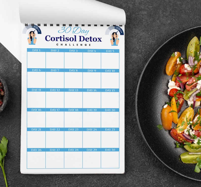 Puede incluir: Un bloc de notas blanco con un borde azul y el texto "30 Day Cortisol Detox Challenge" se muestra. El bloc de notas est&aacute; abierto a un dise&ntilde;o de calendario con d&iacute;as numerados. Un plato negro con una colorida ensalada est&aacute; a la derecha.
