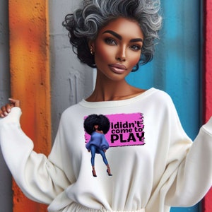 May include: Cream-coloured sweatshirt with a graphic that reads "I didn't come to PLAY." The graphic features a woman in a blue top and jeans. The background is a pink rectangle. The model in the image has grey hair.