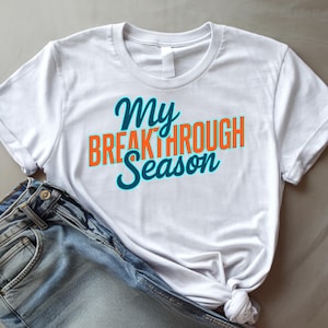 May include: White t-shirt with the text "My Breakthrough Season" in teal and orange. The shirt is paired with blue denim jeans. The text is in a script font.
